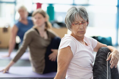 Frauen im Yoga-Kurs, gemischte Altersgruppe, auf Matte sitzend, sich umdrehen, Fokus auf ältere Frau im Vordergrund